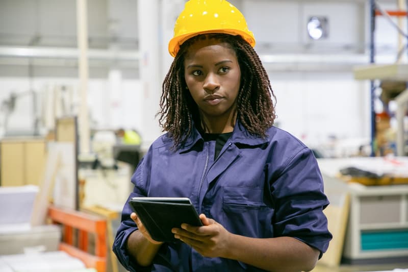 Une opératrice en casque et bleu de travail, tablette en main, dans un atelier.