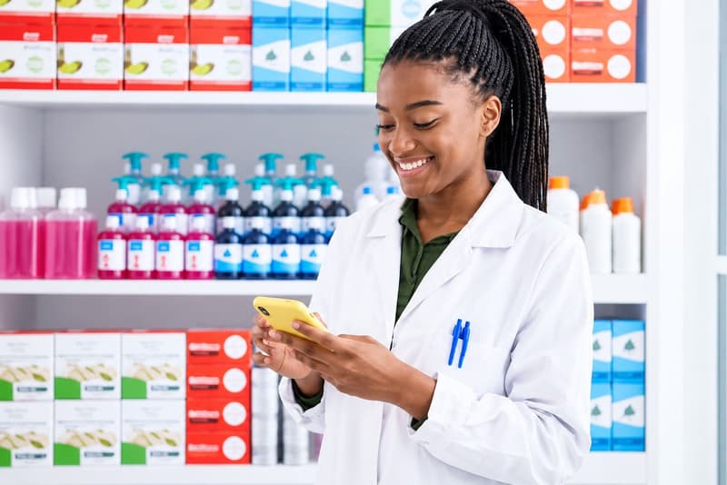 Une pharmacienne en blouse blanche consulte son téléphone devant les rayons de son officine.